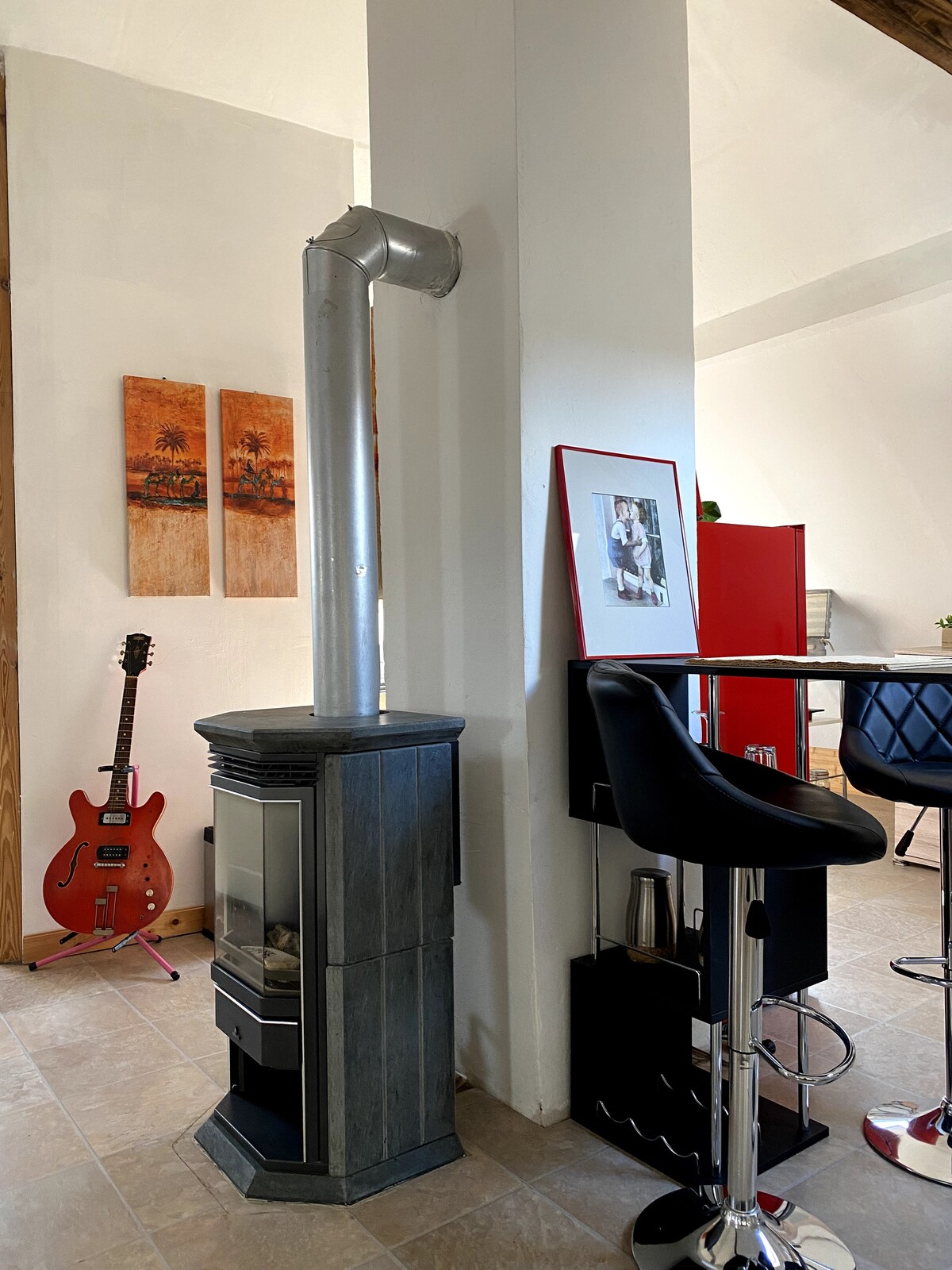A compact space features a sleek gray stove with a metallic flue extending upward. Wall art with two framed images hangs nearby. A vibrant red guitar leans against the wall, while a black bar stool complements the cozy atmosphere of the adjoining area.