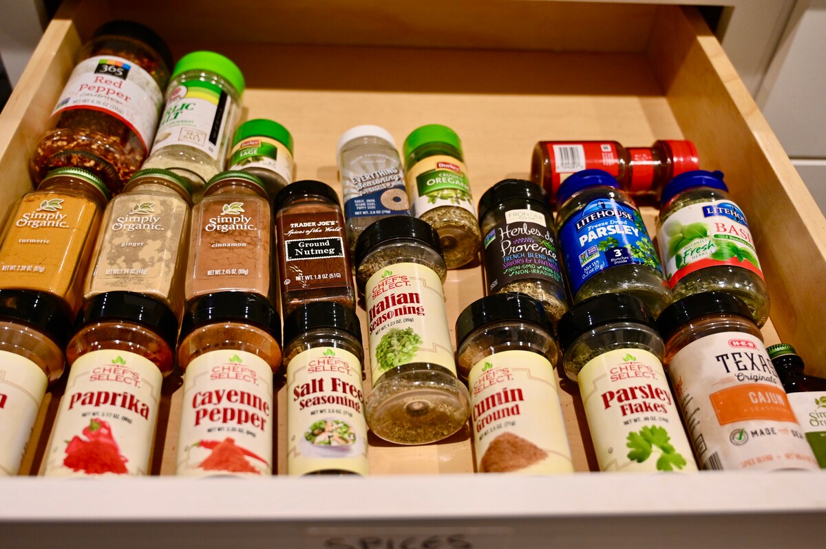 An organized drawer displays an array of spice jars, featuring various labels and colors. Included are spices such as paprika, cayenne pepper, and cumin, alongside smaller containers of seasonings. The arrangement offers easy visibility for cooking needs.