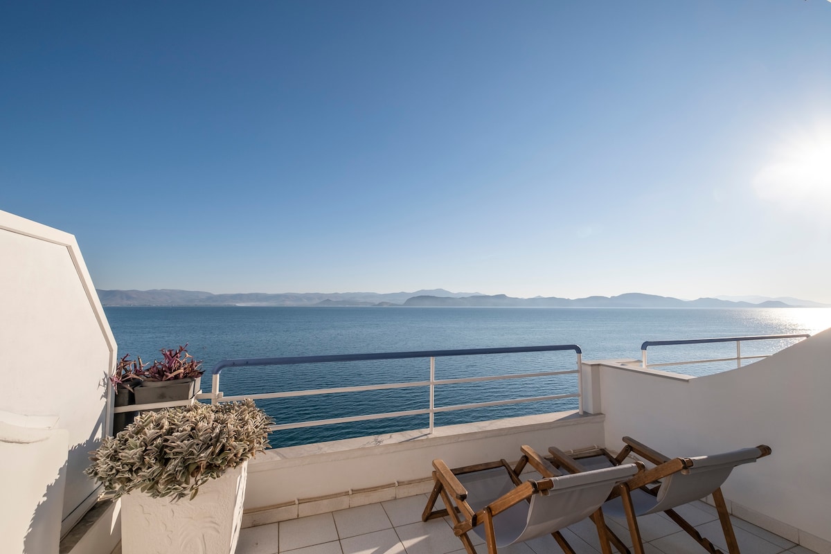 A balcony offers panoramic views of the sea, framed by the expansive sky and distant mountains. Two wooden chairs are positioned to provide a relaxed seating area, while a decorative pot with greenery adds a touch of nature to the space.