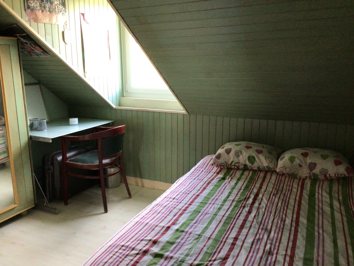 A cozy bedroom features slanted ceilings with green paneling. A spacious bed with striped bedding is positioned alongside a small desk and a chair. A window allows natural light to fill the room, creating a serene and comfortable space.
