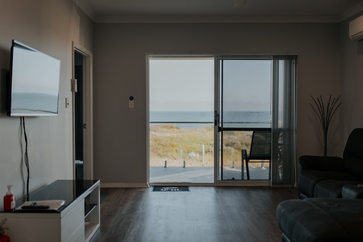 A spacious living area is visible, featuring a large glass door that opens to an unobstructed view of the ocean. A flat-screen television is mounted on the wall, while comfortable seating arrangements invite relaxation. Natural light streams in from the doorway, enhancing the calming atmosphere.