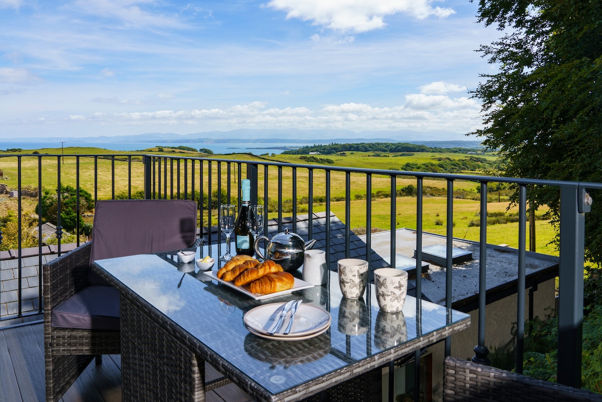 A dining table is set on a balcony, featuring plates with croissants, two mugs, and a wine bottle. Comfortable seating is provided by two armchairs. A scenic view of grassy fields and the distant coastline can be seen in the background under a partly cloudy sky.