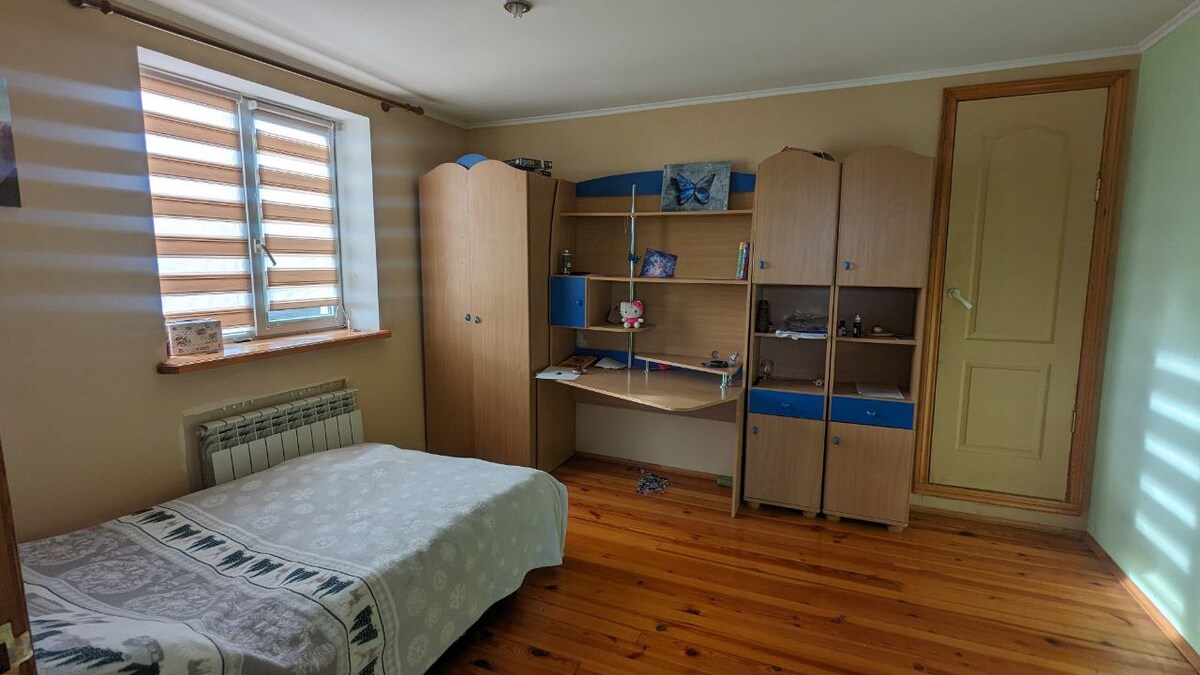 A neatly arranged bedroom features a spacious bed with a patterned coverlet, accompanied by wooden furniture including a wardrobe and a desk. Natural light enters through a window with horizontal blinds, casting subtle stripes across the room's light-colored walls and wooden flooring.