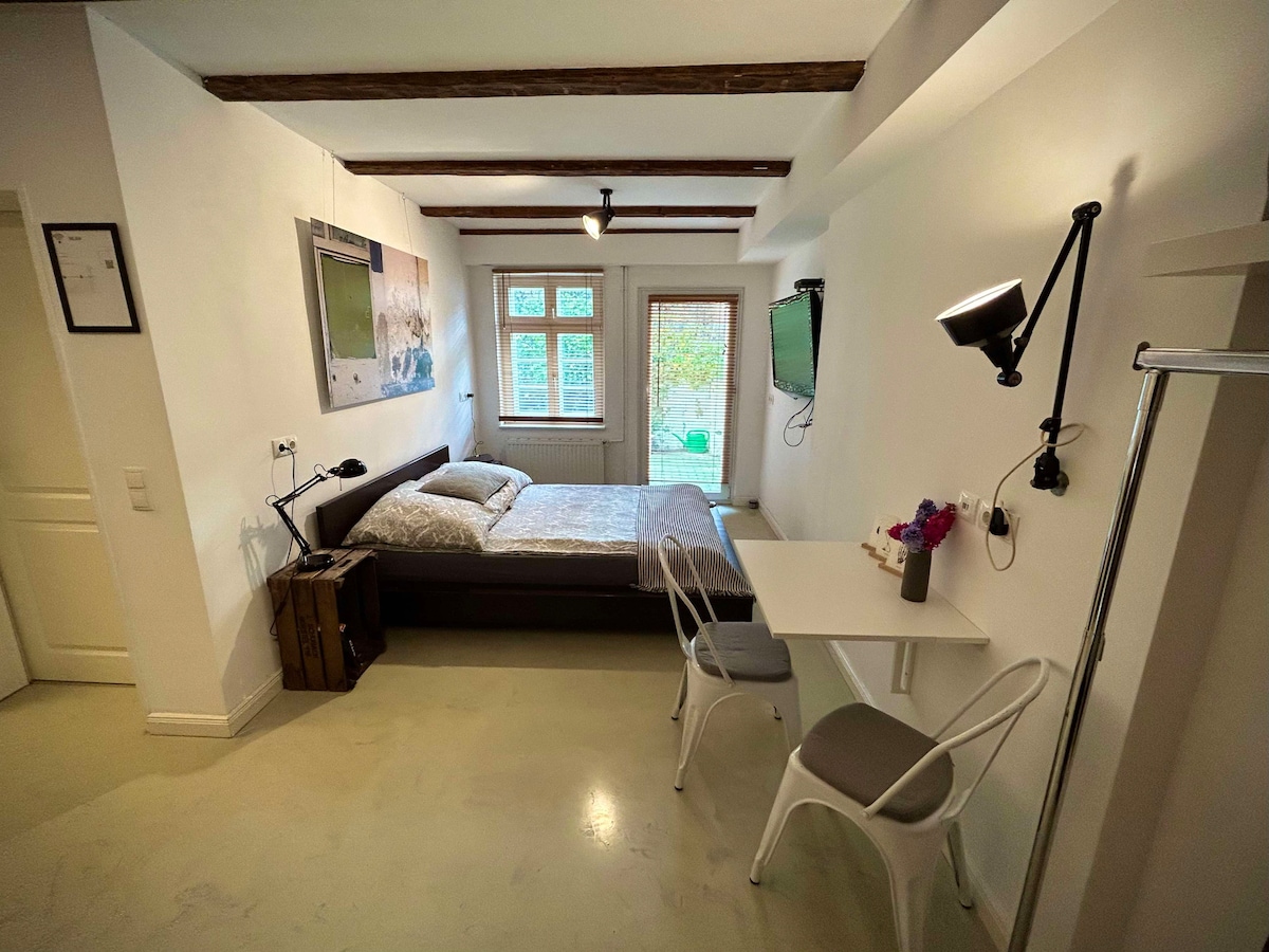 A well-furnished room features a spacious bed with neutral bedding, complemented by natural light from a window. A small dining table with two chairs is positioned nearby. A flat-screen TV is mounted on the wall, and a door leads to a small outdoor space.