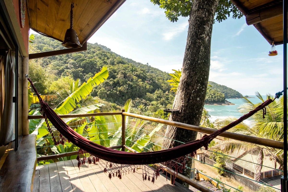 A balcony space features a hammock suspended between wooden posts, overlooking a lush green landscape and the ocean in the distance. The scene is framed by tropical foliage, allowing natural light to fill the area with a serene ambiance.