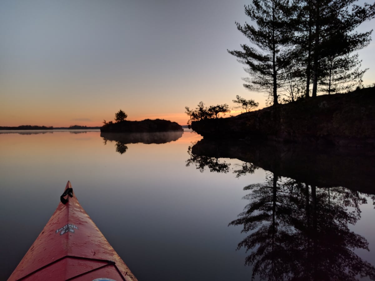 A kayak is positioned at the forefront of a calm lake, reflecting the surrounding trees and the soft hues of a sunset. The still water creates a serene atmosphere, with gentle ripples appearing near the rocky shorelines.