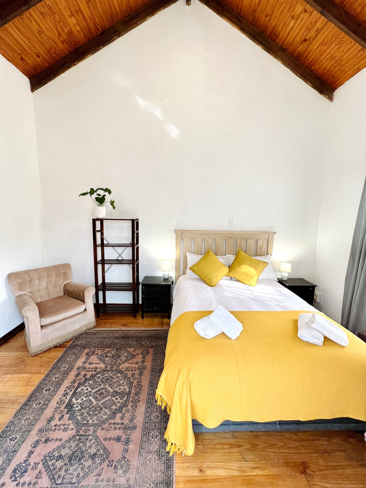 A cozy room features a spacious bed adorned with yellow and white linens. Flanking the bed are two nightstands with lamps, alongside a decorative area rug. A comfortable chair is positioned in one corner, and natural light filters through a window with grey curtains.