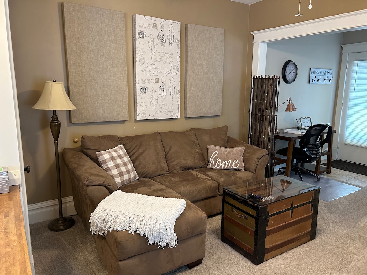 A comfortable living area showcases a sectional sofa in a warm brown hue, accompanied by a decorative throw. Neutral wall art is displayed above, while a glass-top coffee table stands in front. A desk and chair are positioned nearby, with natural light filtering through a window.