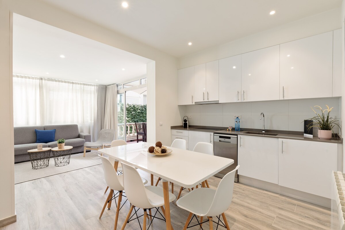 The modern kitchen features sleek white cabinetry and appliances, complemented by a light wood dining table surrounded by white chairs. Natural light fills the open layout, revealing a comfortable living area with a gray sofa, a coffee table, and a terrace with greenery visible through glass doors.