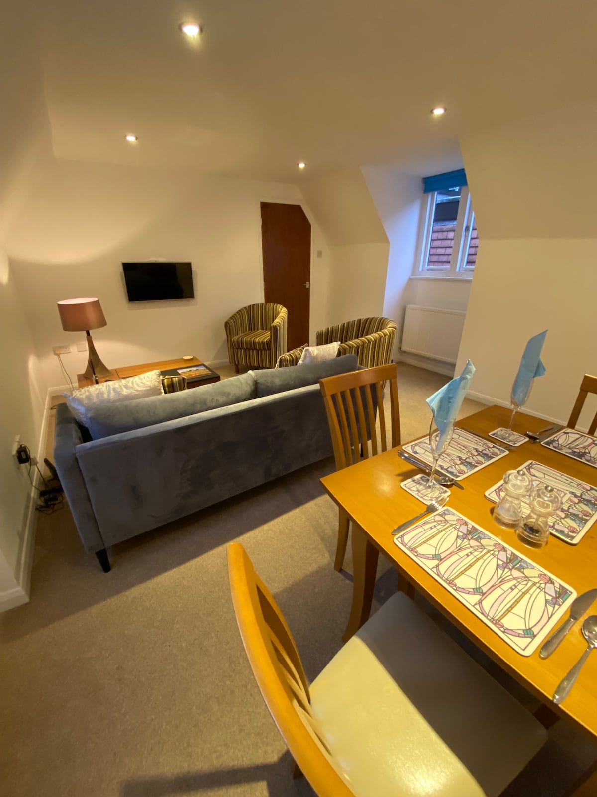 A cozy lounge area is furnished with a plush, light blue sofa and two striped armchairs. A wooden dining table is set with blue and white accents, accommodating four people. Soft lighting creates a welcoming ambiance, and a flat-screen TV is mounted on the wall.