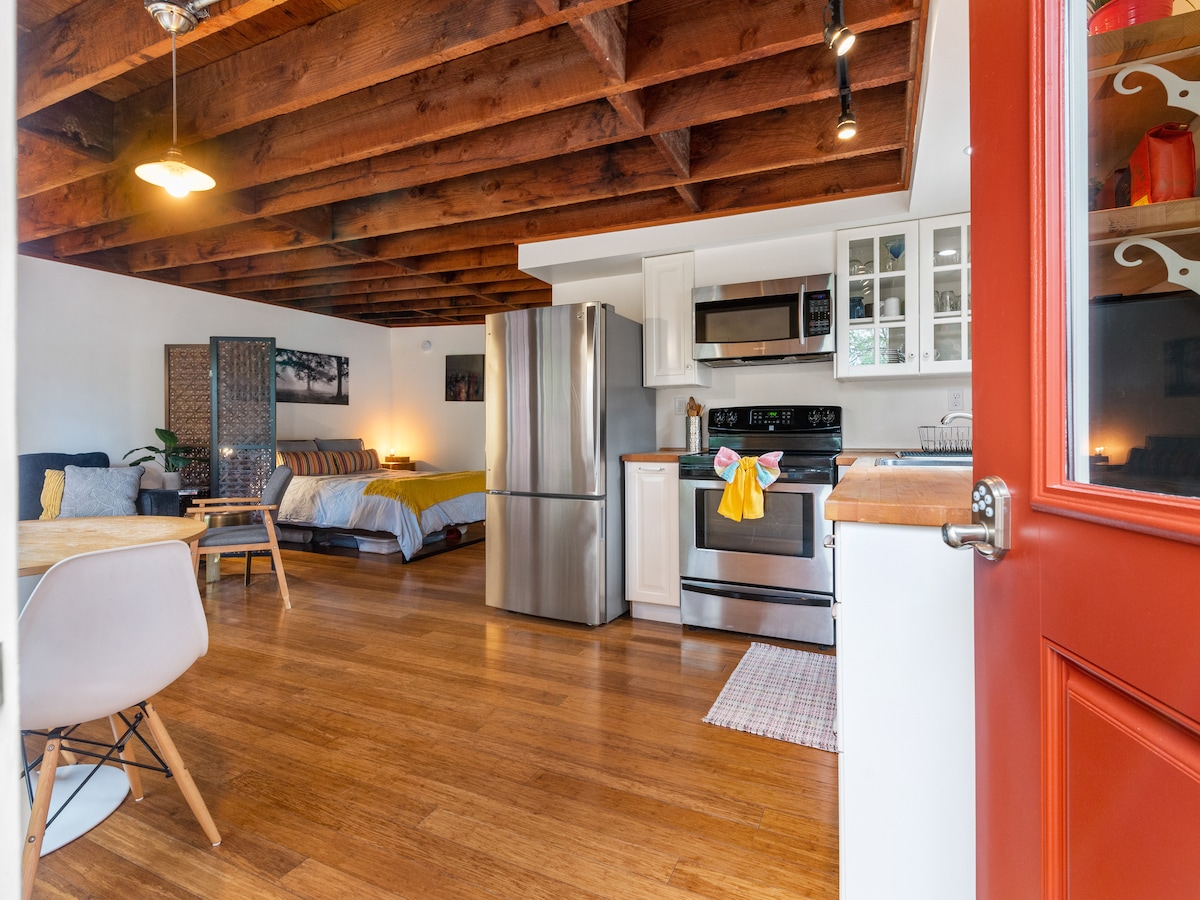 The open living space features wood beams overhead, a fully equipped kitchen with stainless steel appliances, and a cozy dining area. A comfortable bed area is visible in the background, accompanied by a sleek sofa and natural light filtering through the windows.