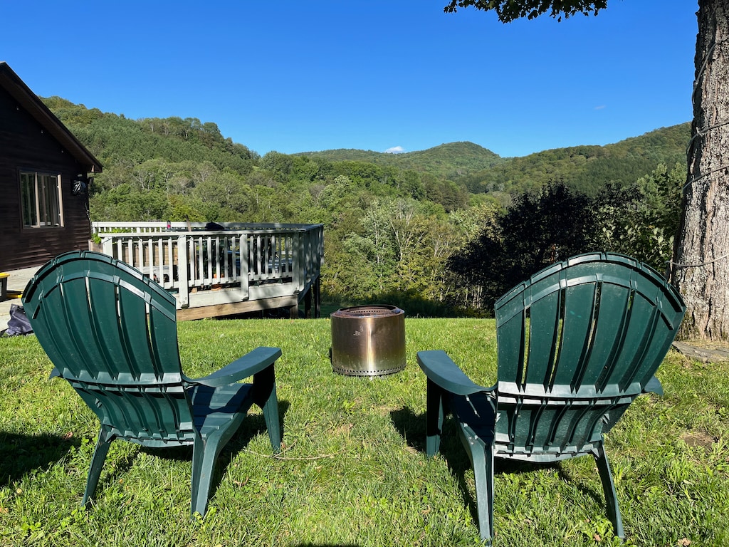 Two green Adirondack chairs are positioned on a grassy lawn, facing a metal fire pit. Lush green hills are visible in the background under a clear blue sky, providing a serene outdoor setting for relaxation.