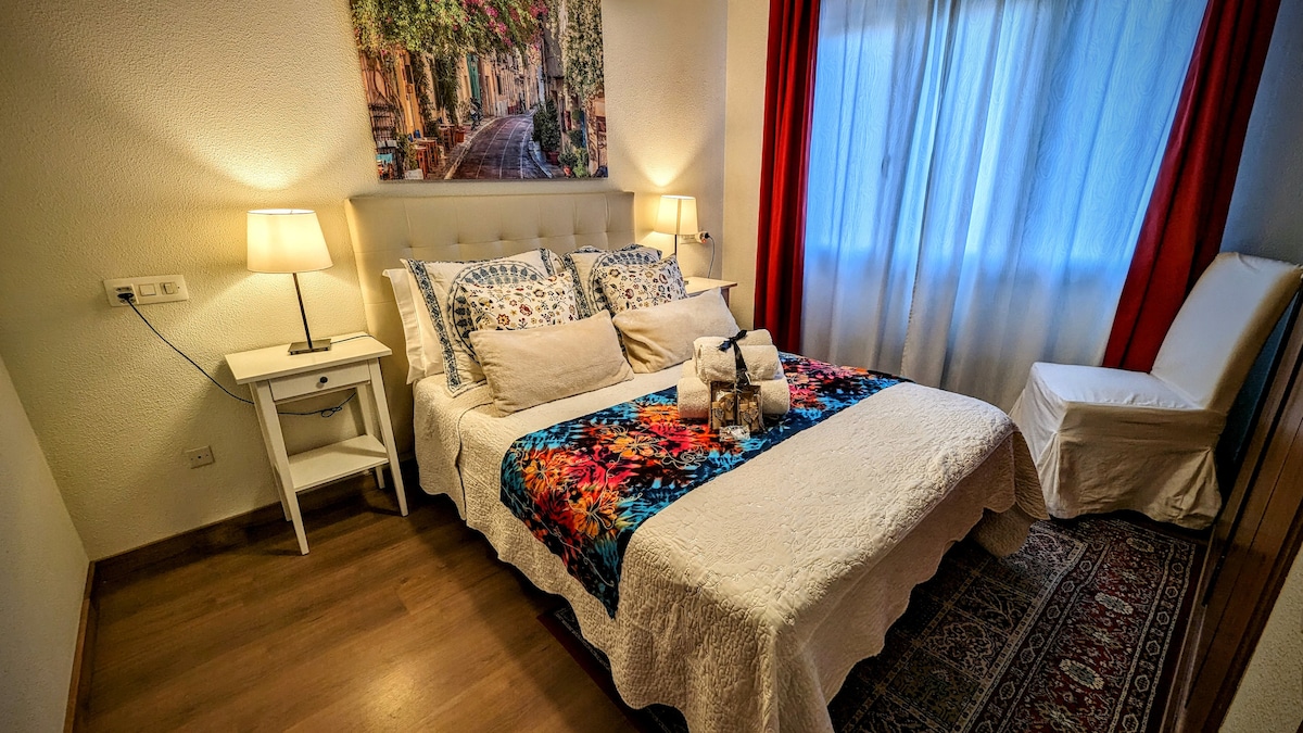 A well-appointed bedroom features a spacious bed adorned with a colorful cover and decorative pillows. Soft lighting is provided by bedside lamps, while natural light filters through sheer curtains. A comfortable chair and a rug add to the room's inviting ambiance.