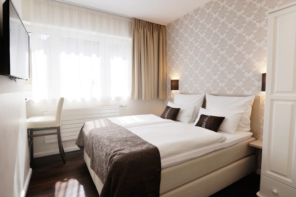 A well-appointed room features a spacious bed with folded linens and decorative cushions. Natural light filters through sheer curtains, illuminating the soft wallpaper. A light-colored chair and small table are positioned nearby, and a flat-screen TV is mounted on the wall.