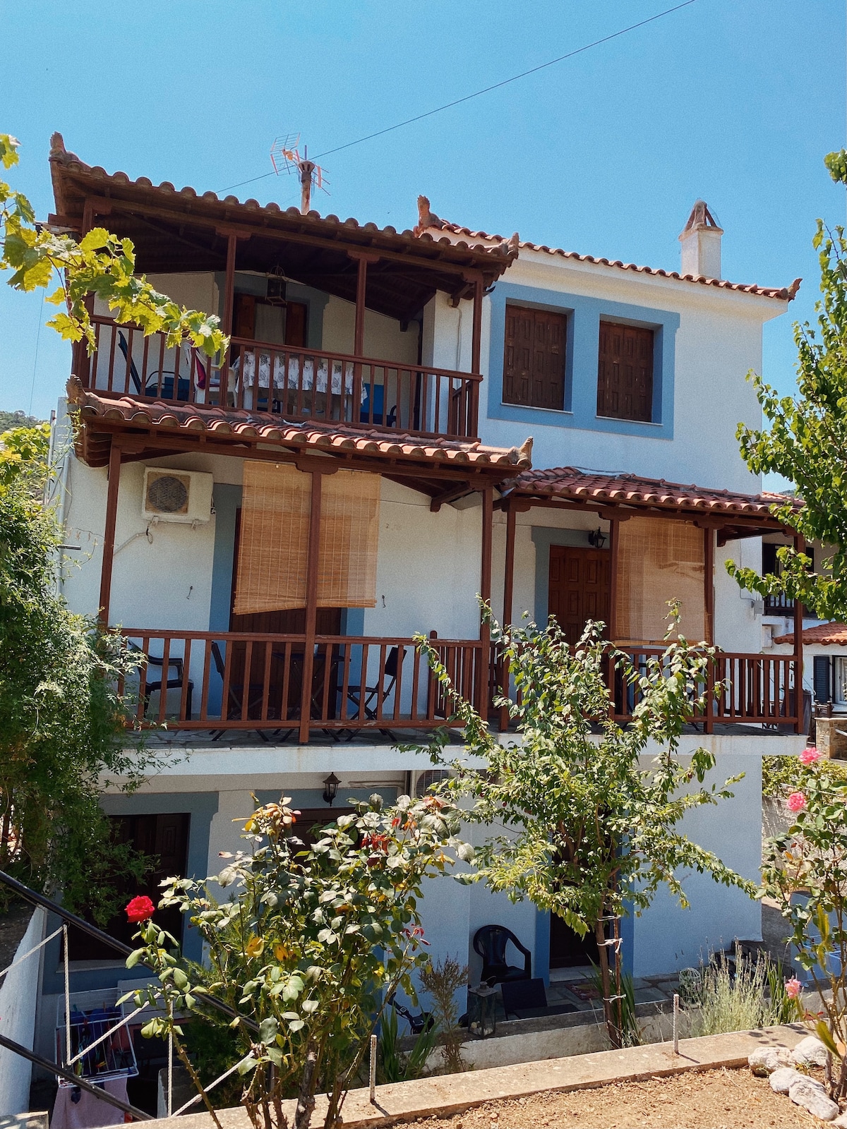 The three-story building is constructed in traditional island architecture, featuring wooden balconies and vibrant blue and white walls. Lush greenery surrounds the property, with blooming flowers and small trees at the forefront, creating a welcoming outdoor space.