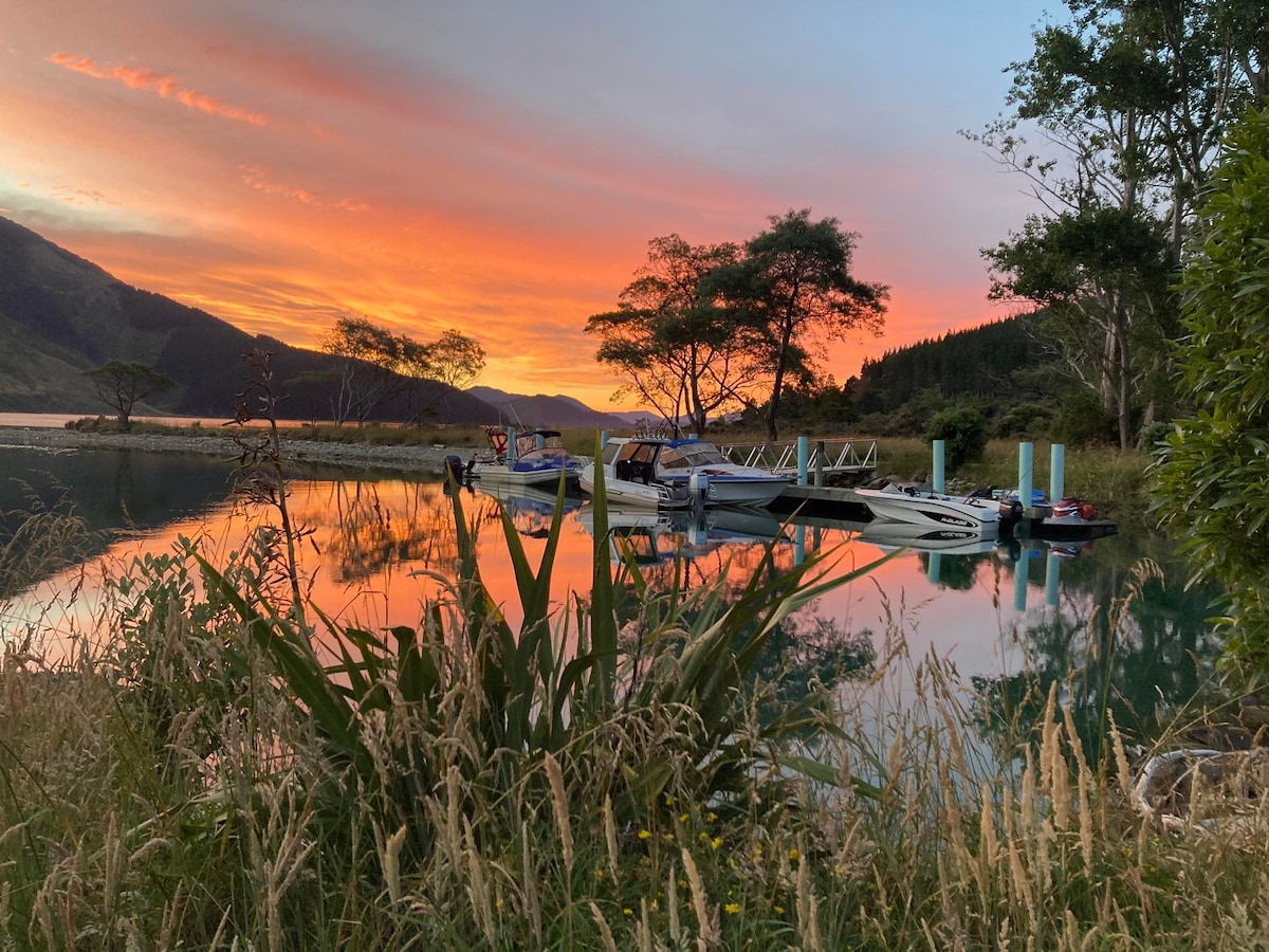 Calm waters reflect a vibrant sunset, surrounded by lush greenery and distant hills. Several boats are moored at a floating jetty, contributing to the serene ambiance of the Marlborough Sounds.