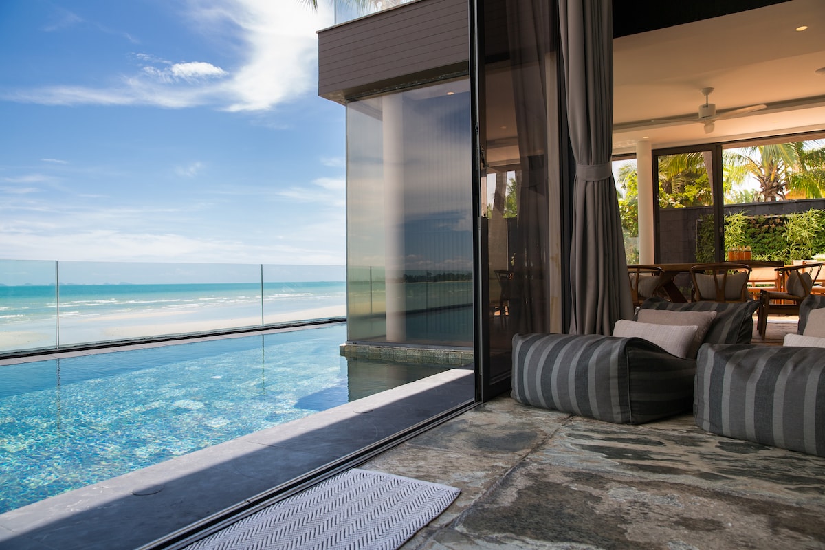 A serene view is presented from the main living area, where a spacious infinity pool mirrors the sky, with the ocean stretching beyond. Comfortable seating is arranged near large glass doors that provide a seamless transition to the outdoor space, enhanced by lush tropical surroundings.