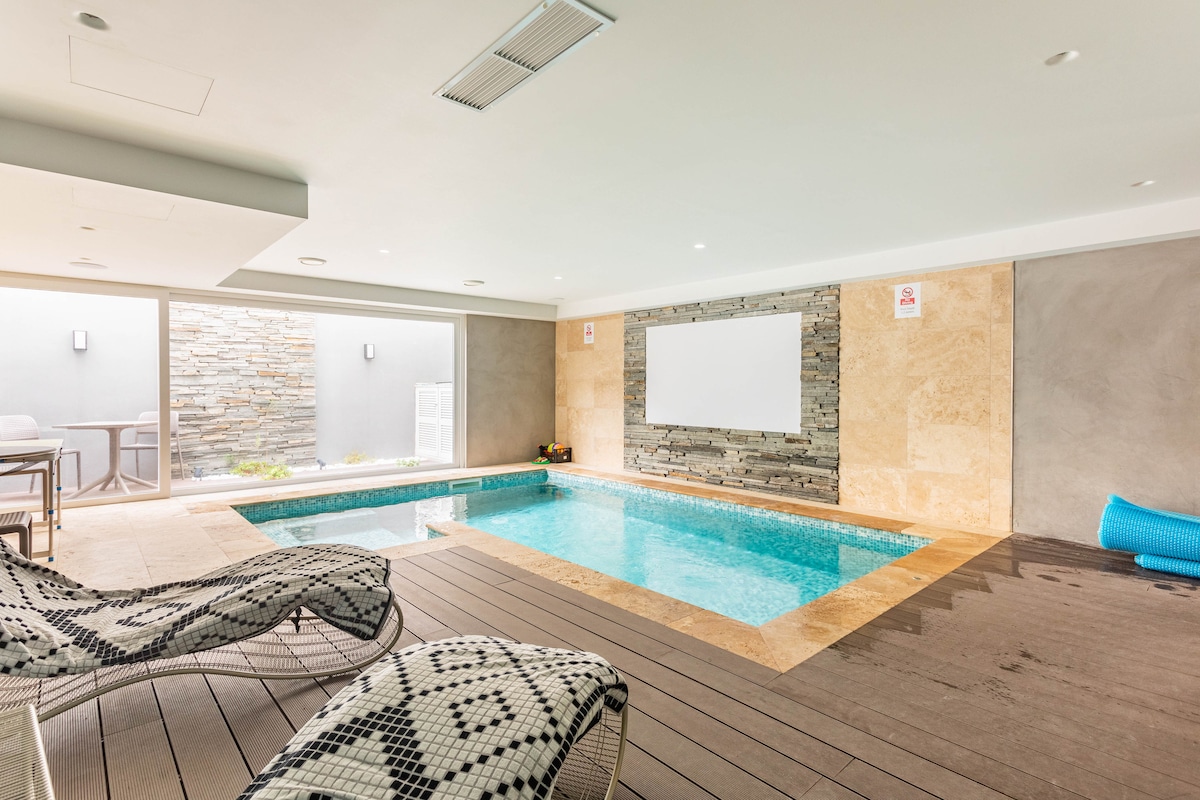 A heated indoor pool area features a smooth surface with decorative tile, complemented by natural stone wall accents. Sun loungers are positioned nearby, creating a space for relaxation. Large glass panels allow natural light to fill the area, enhancing the cheerful ambiance.