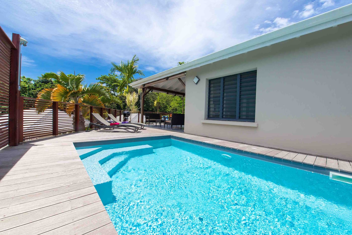 A private pool is surrounded by a wooden deck, with lounge chairs positioned for relaxation. A shaded terrace is visible in the background, offering seating for outdoor dining. Lush greenery and palm trees frame the scene, enhancing the villa's tranquil atmosphere.