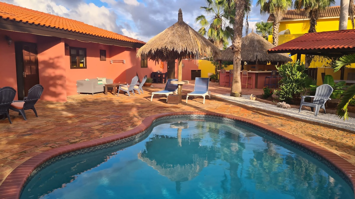 A swimming pool is framed by a tiled patio, featuring several lounge chairs and a shaded palapa. Colorful casitas with red and yellow walls are visible in the background, surrounded by tropical landscaping and palm trees, creating a tranquil outdoor environment.