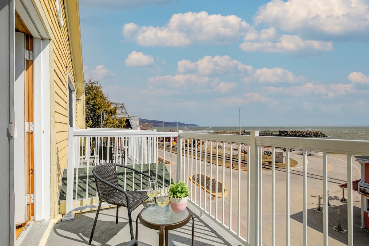A balcony is seen with a small round table and a chair, offering a view of the river and surrounding landscape. The soft colors of the exterior walls complement the bright sky, creating a serene outdoor space suitable for relaxation.