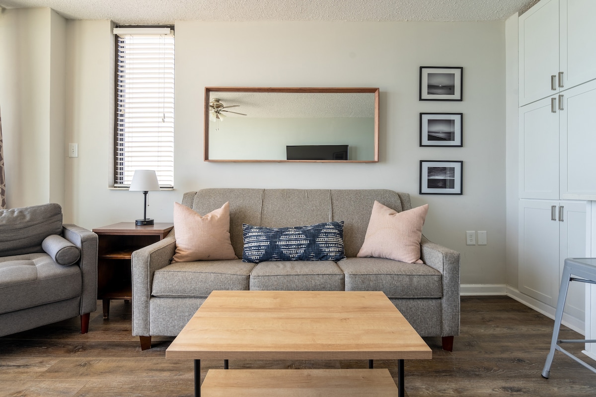 A modern living room features a comfortable gray sofa adorned with decorative pillows. A wooden coffee table is positioned in front of the sofa. A large mirror reflects the light coming from the window. Photographs are displayed on the wall, alongside a compact side table with a lamp.