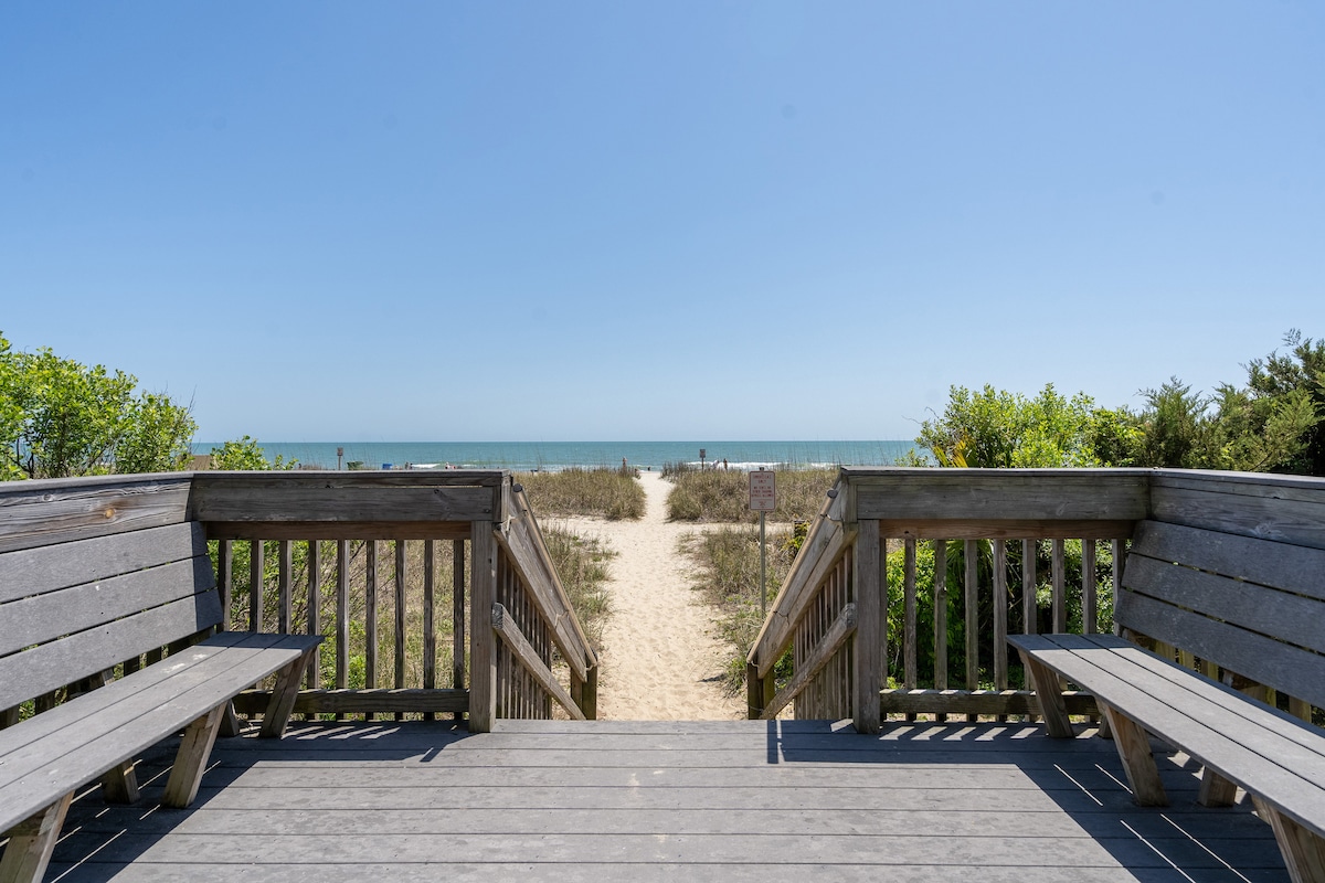 A wooden boardwalk leads to the sandy beach, flanked by lush greenery on both sides. The sunlit ocean is visible in the distance, with gentle waves lapping the shore. The path is clear, inviting guests to enjoy the seaside views.