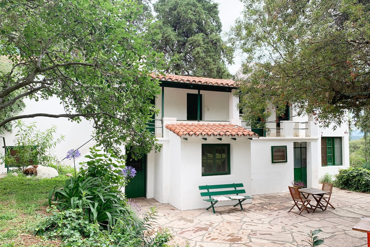 A charming exterior of the studio displays a traditional design with a terracotta roof and green window shutters. Surrounding gardens feature lush greenery and vibrant flowers. A wooden dining table and chairs are positioned on the stone patio, offering a tranquil outdoor space.