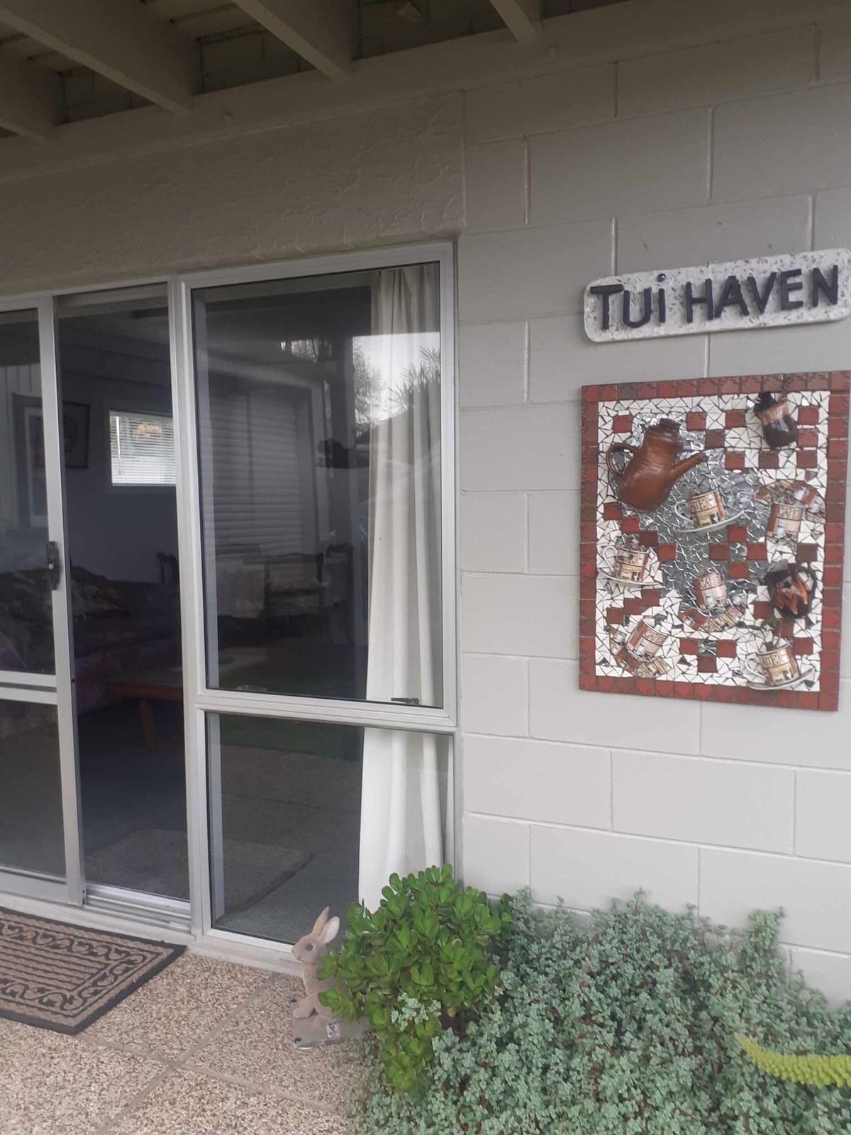 An entrance to Tui Haven is shown, featuring sliding glass doors leading into the unit. A decorative mosaic sign displaying 'Tui Haven' is mounted on the wall beside the entrance. A small garden area with greenery is visible in the foreground.