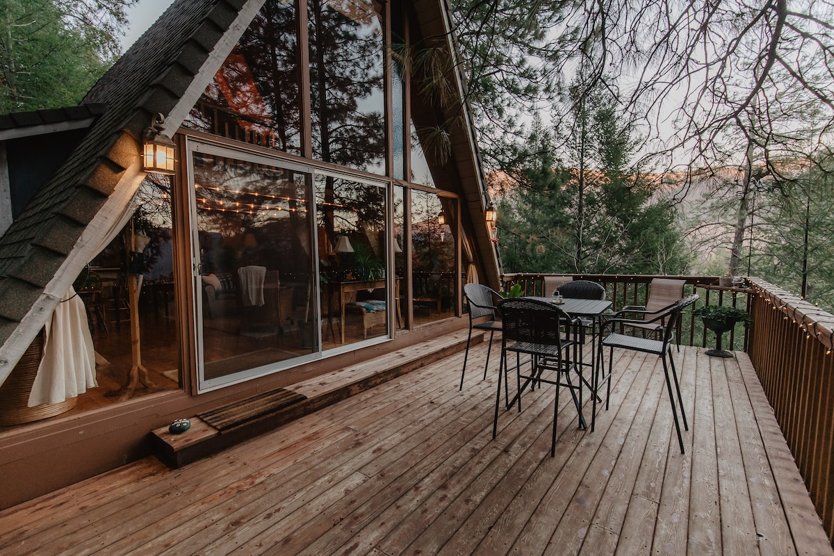 A spacious front deck features a wooden surface surrounded by trees, showcasing a glass sliding door that reflects the natural surroundings. A dining set with four chairs and a table is positioned for outdoor enjoyment, inviting guests to relax in the serene environment.