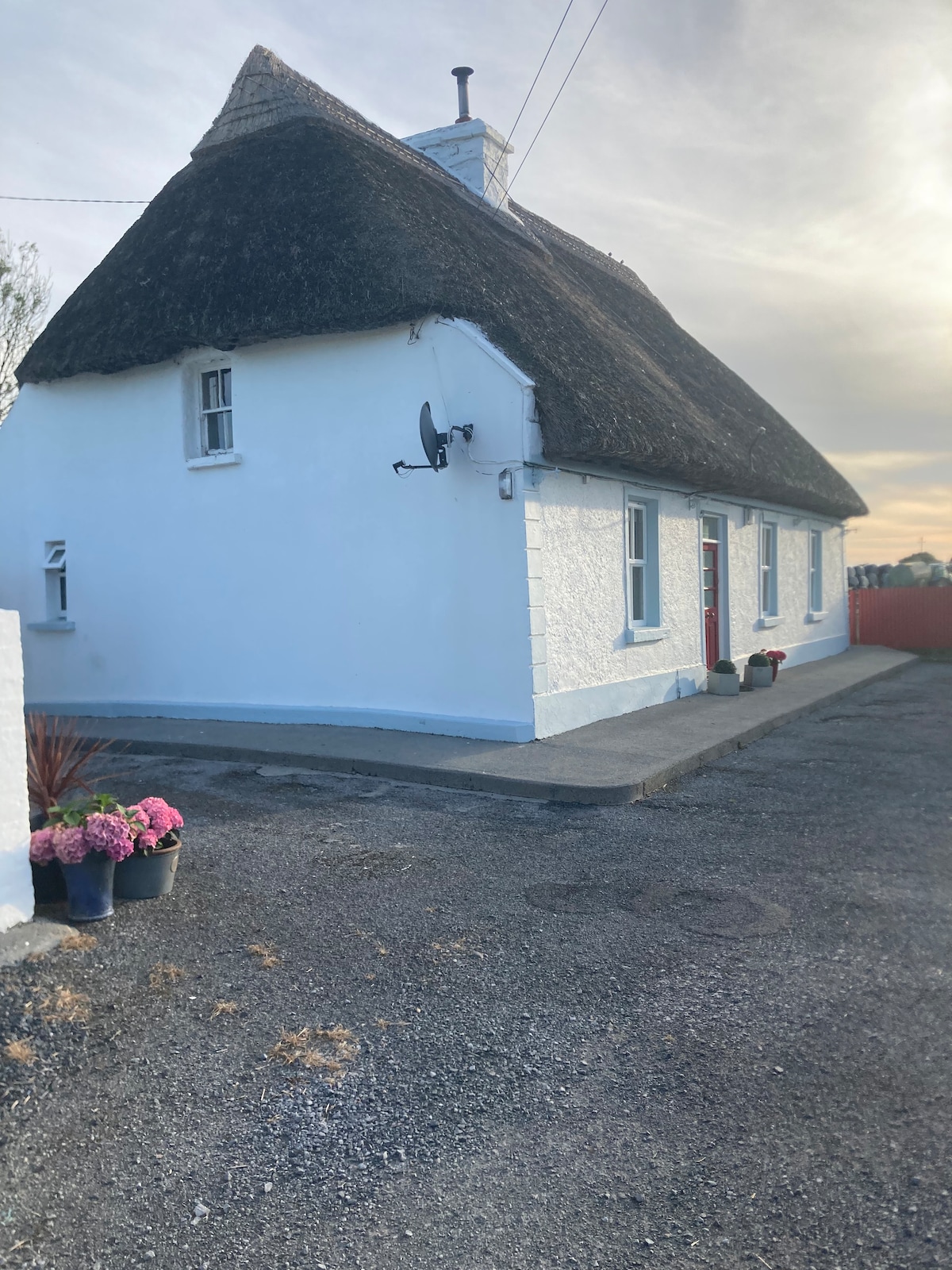 The charming cottage features a thatched roof and whitewashed walls, set against a serene sky. Flowers in pots are positioned along the entrance, contributing to the welcoming outdoor space. A gravel driveway leads to the front door of the cottage.