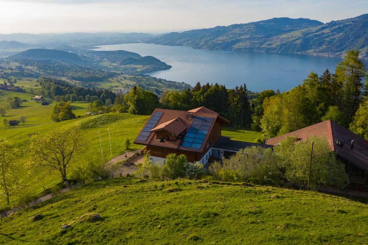 A panoramic view captures a hillside retreat surrounded by lush green fields. Solar panels reflect sunlight on the modern building, with the serene Thunersee stretched out below amidst rolling hills and distant mountains.