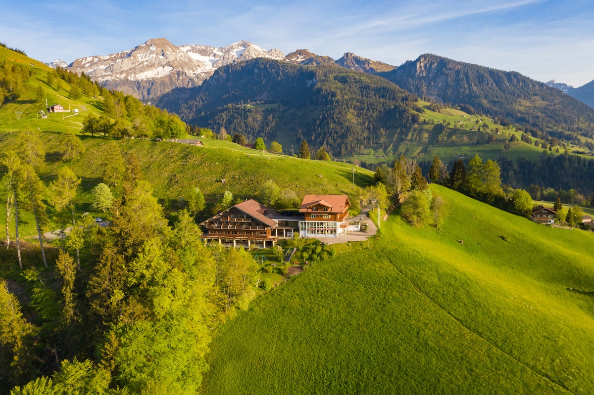 Aerial view of a modern chalet-style hotel nestled among rolling green hills. Snow-capped mountains rise in the background, while trees and meadows surround the property, creating a serene landscape.