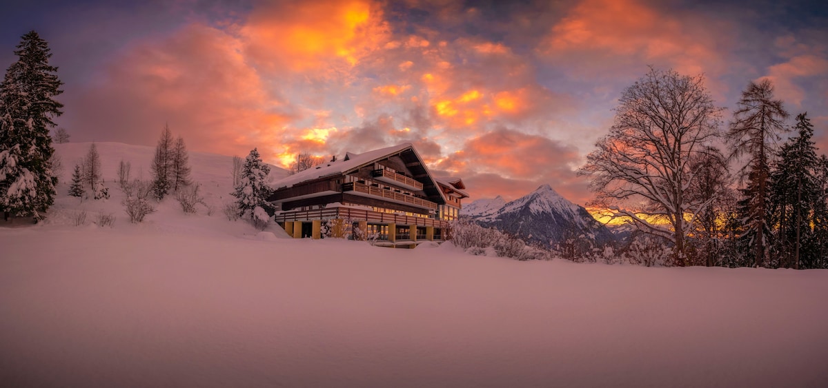 The chalet is surrounded by a blanket of fresh snow, reflecting the warm hues of a sunset sky. Majestic mountains rise in the background, contributing to the serene winter landscape. The building features numerous windows, allowing for natural light to fill its interior.
