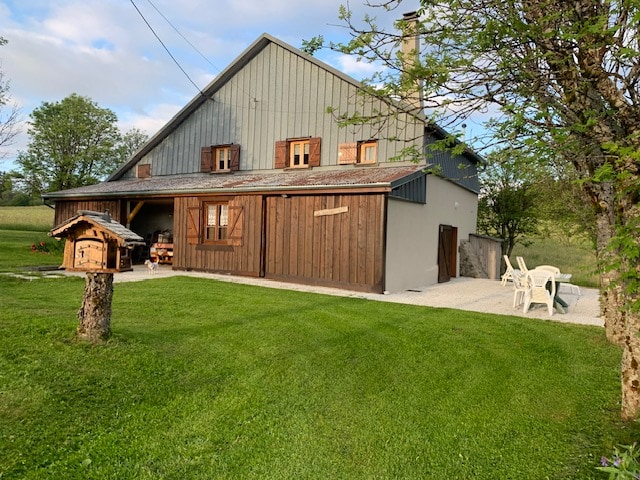 The exterior of the chalet is displayed, featuring a combination of wooden and grey walls with multiple shuttered windows. A small birdhouse is visible in the foreground, along with a set of four white chairs positioned on a gravel area. Lush green grass surrounds the property.