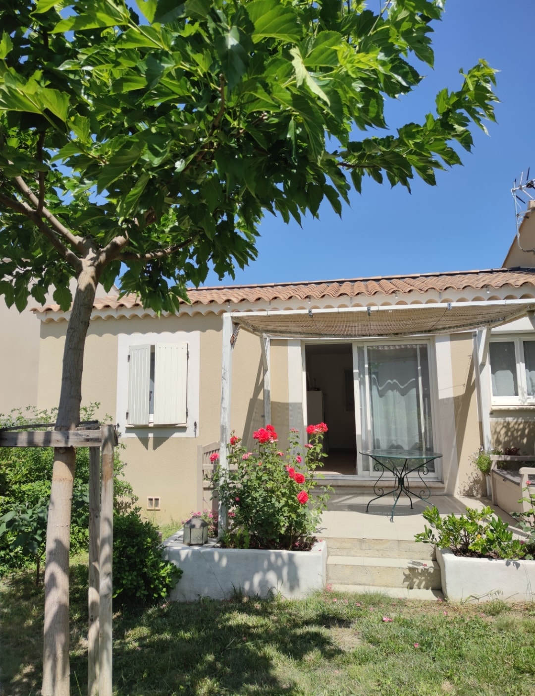 The exterior view of the house features a covered terrace with a small table and chairs. Green foliage and blooming flowers surround the entrance, and the sunny sky highlights the peaceful ambiance of the outdoor space.