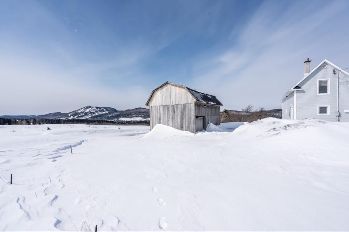 A rustic wooden shed is positioned amongst a landscape blanketed in snow, with gentle hills visible in the distance. The exterior of a light-colored house provides contrast against the winter scenery, showcasing the serene atmosphere of the countryside.