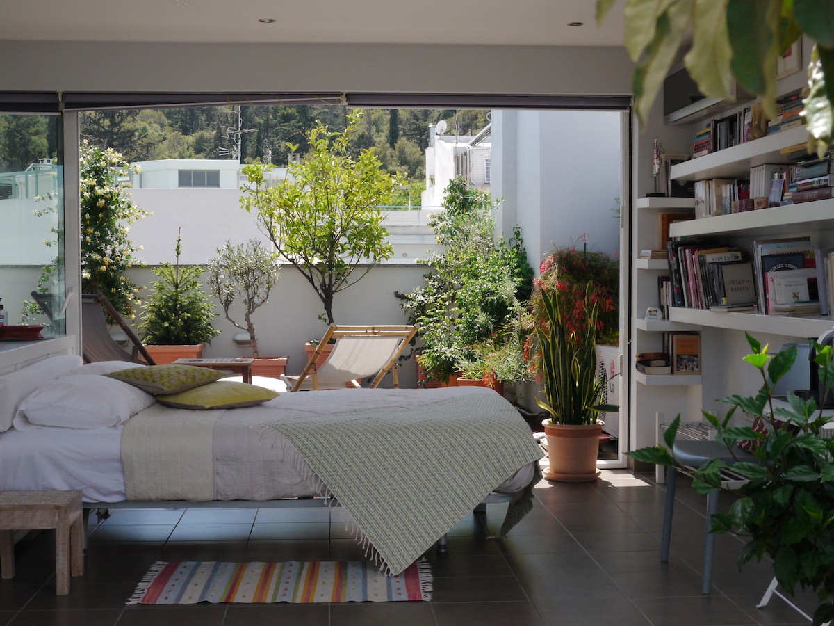 A bright and airy loft apartment features a spacious bed with a soft blanket. A private terrace is visible through large glass doors, framed by greenery and potted plants. Sunlight enhances the serene atmosphere, while a cozy reading nook with shelves is present in the background.