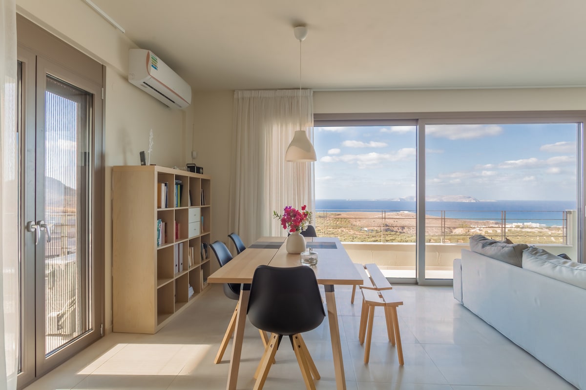 A bright and airy living space features a long dining table surrounded by black chairs. A light-colored bookshelf stands nearby, with a vase of flowers in the center of the table. Expansive windows reveal a scenic view of the coastline.
