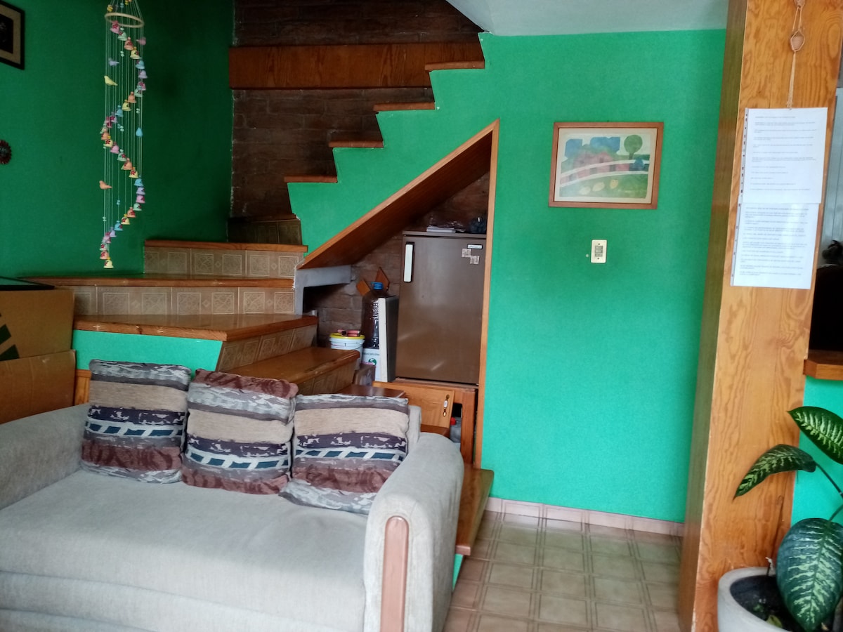A cozy living area features a light brown sofa adorned with decorative pillows. A green wall contrasts with the wooden stairs leading to the upper level. A small refrigerator is partially visible in the background, adding functionality to the space.