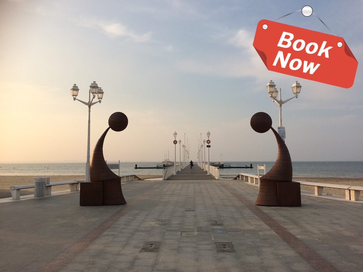 The image captures a scenic pier extending toward the horizon, framed by smooth waves and a soft pastel sky. Two abstract sculptures are positioned on either side of the walkway, while lampposts line the path, guiding the way to the beach.