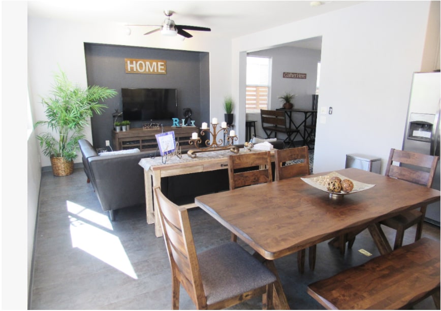 A welcoming living area combined with a dining space features a wooden table set for six. The cozy sofa faces a mounted television, surrounded by decor and plants. Natural light fills the room through nearby windows, enhancing the minimalist design.