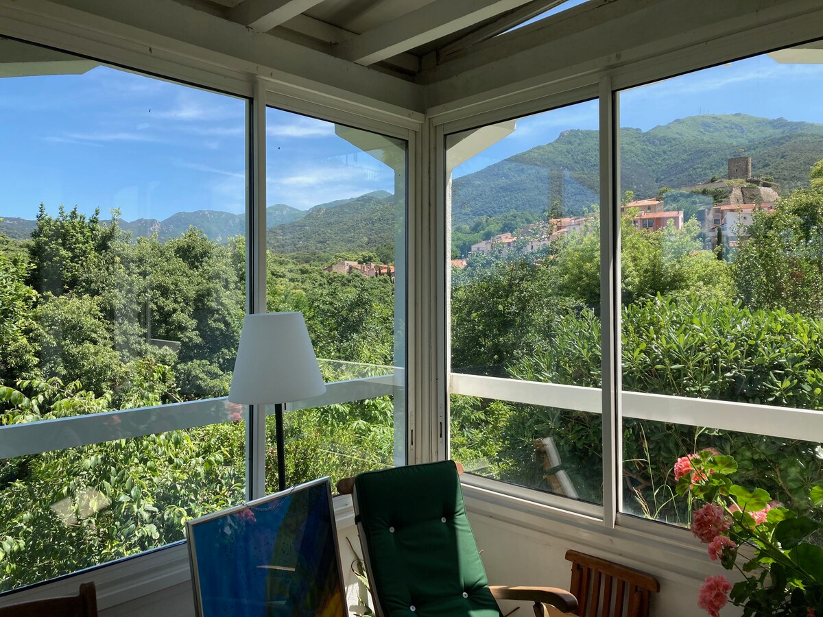 A sunlit conservatory is adorned with large glass windows, offering panoramic views of lush greenery, rolling hills, and distant mountains. A comfortable chair is positioned nearby, alongside a lamp and a small table with an art piece, creating a tranquil corner to enjoy the scenery.