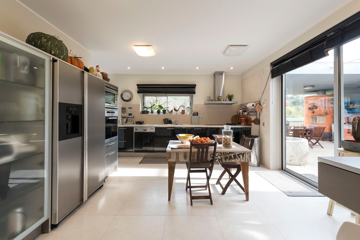 The modern kitchen features sleek black cabinetry and stainless steel appliances, including a large refrigerator. A wooden dining table is set with fruit, surrounded by chairs. Natural light fills the space through a large window leading to an outdoor dining area.