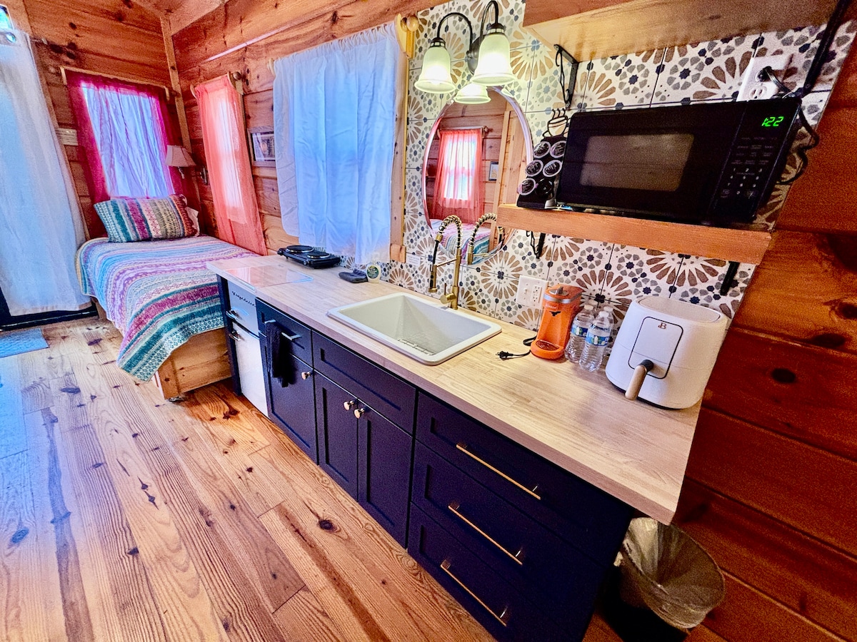 The kitchenette features a wood countertop with a dual sink, a microwave, and a coffee maker. A vibrant patterned backsplash provides visual interest. A colorful bedspread is visible in the background, complemented by sheer window treatments that allow natural light to filter through.