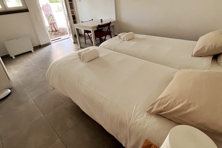 Quarto no anexo exterior, com WC privado e possibilidade de até 4 camas individuais. Duas camas montadas, roupa clara, luz natural e acesso direto ao exterior.