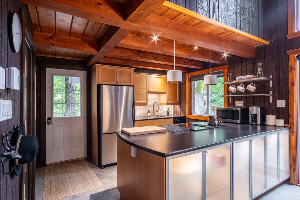 A modern kitchen is highlighted with sleek appliances and wooden cabinetry. The spacious island features a countertop with a cutting board, while pendant lights offer illumination. Large windows provide natural light, enhancing the welcoming atmosphere of the space.