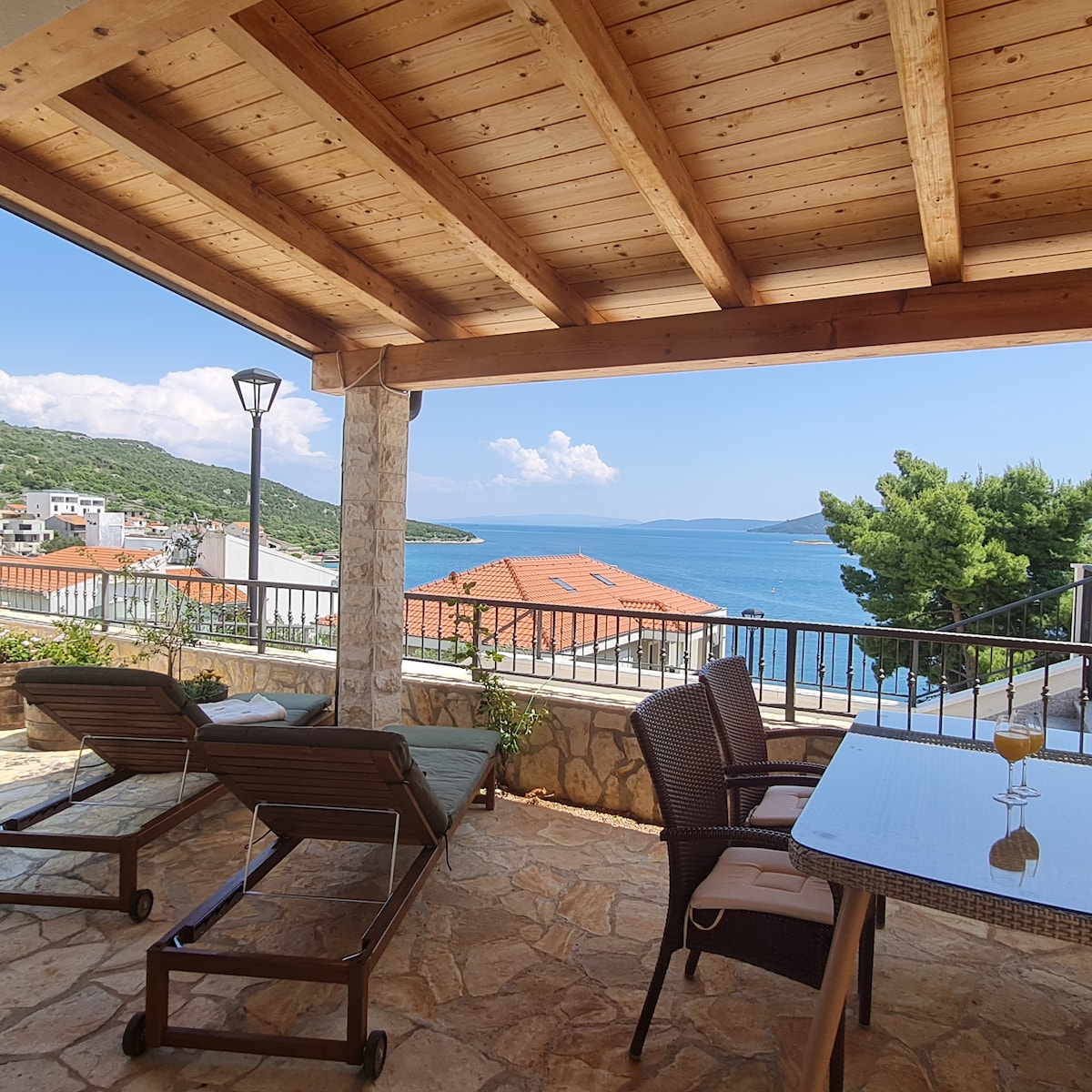 An outdoor terrace is furnished with lounge chairs and a dining table, overlooking a scenic view of the sea and surrounding landscape. A wooden ceiling adds warmth to the space, while a stone railing provides a sense of openness and connection to the natural surroundings.