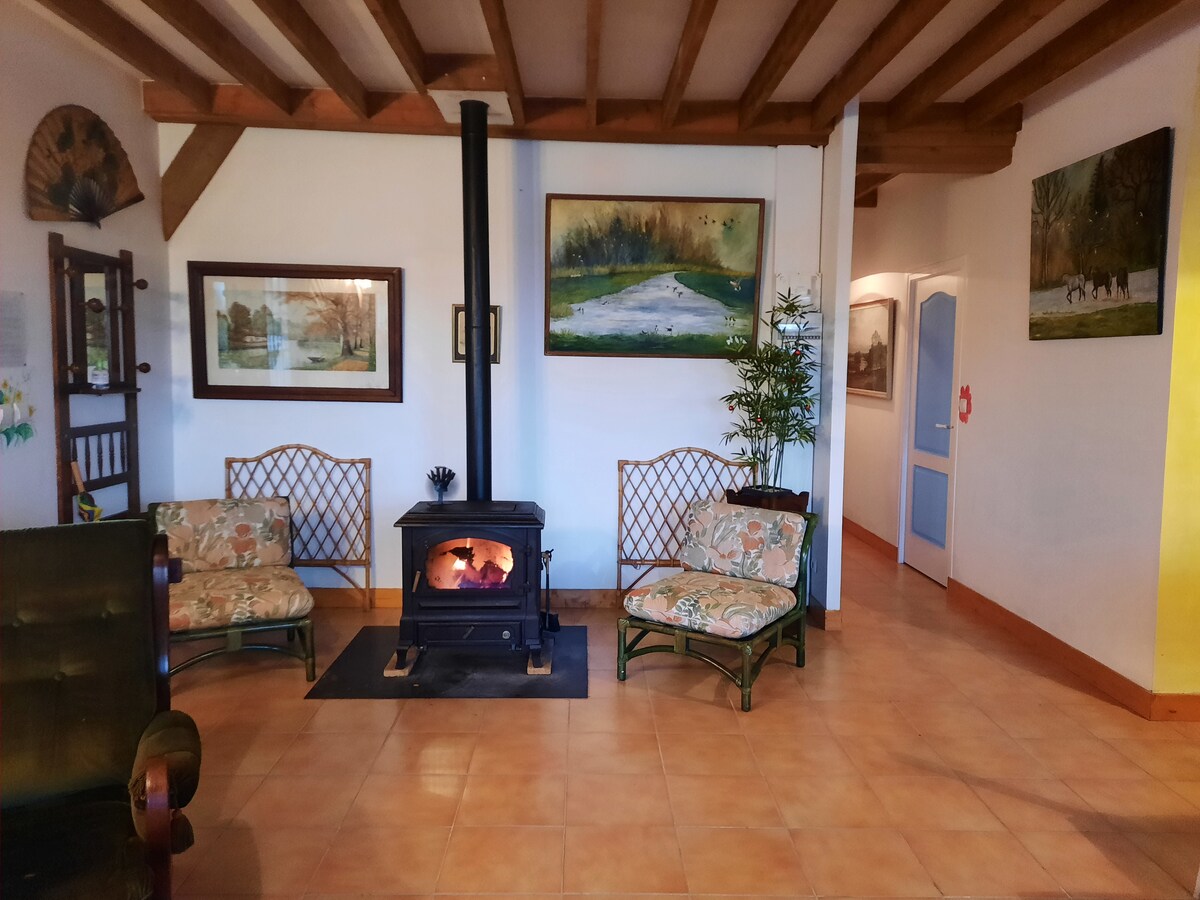 The living area features two comfortable seating arrangements with floral-patterned cushions. A charming wood stove is centrally positioned, enhancing the space. Artistic paintings adorn the walls, contributing to a serene atmosphere. Exposed wooden beams add character to the ceiling.