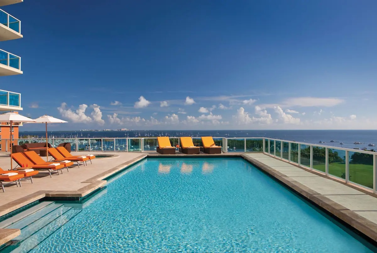 A spacious pool deck is featured with a clear blue pool and inviting lounge chairs positioned under a bright sky. The expansive ocean view is visible in the background, enhancing the serene atmosphere of the outdoor space.